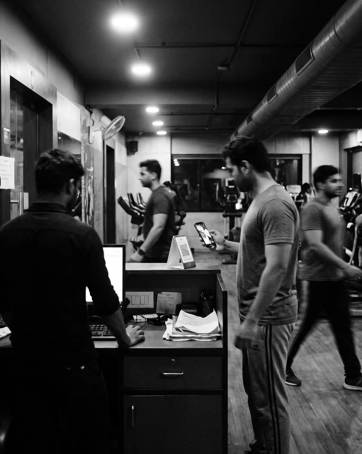 A gym front desk on a weekday evening, a member holding up a phone to the QR check-in stand